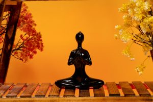 Black figurine sitting cross-legged with hands in prayer position on an orange background, symbolising grounding, mindfulness, and calm.