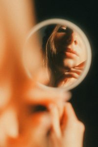 Blurred figure of a woman with her face clearly reflected in a mirror, symbolising self-reflection, awareness, and noticing inner patterns.